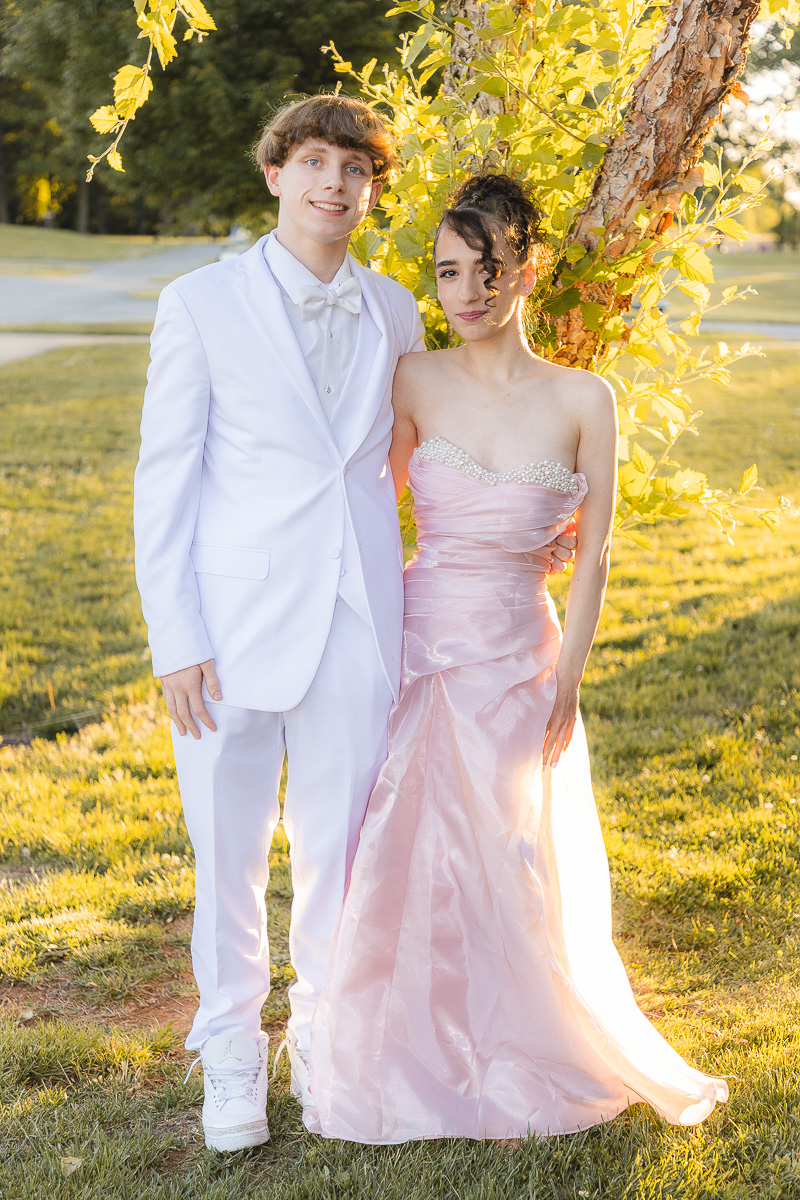 Young couple in formal wear posing outdoors by a tree in a sunlit park (boy in white suit, girl in pink gown).