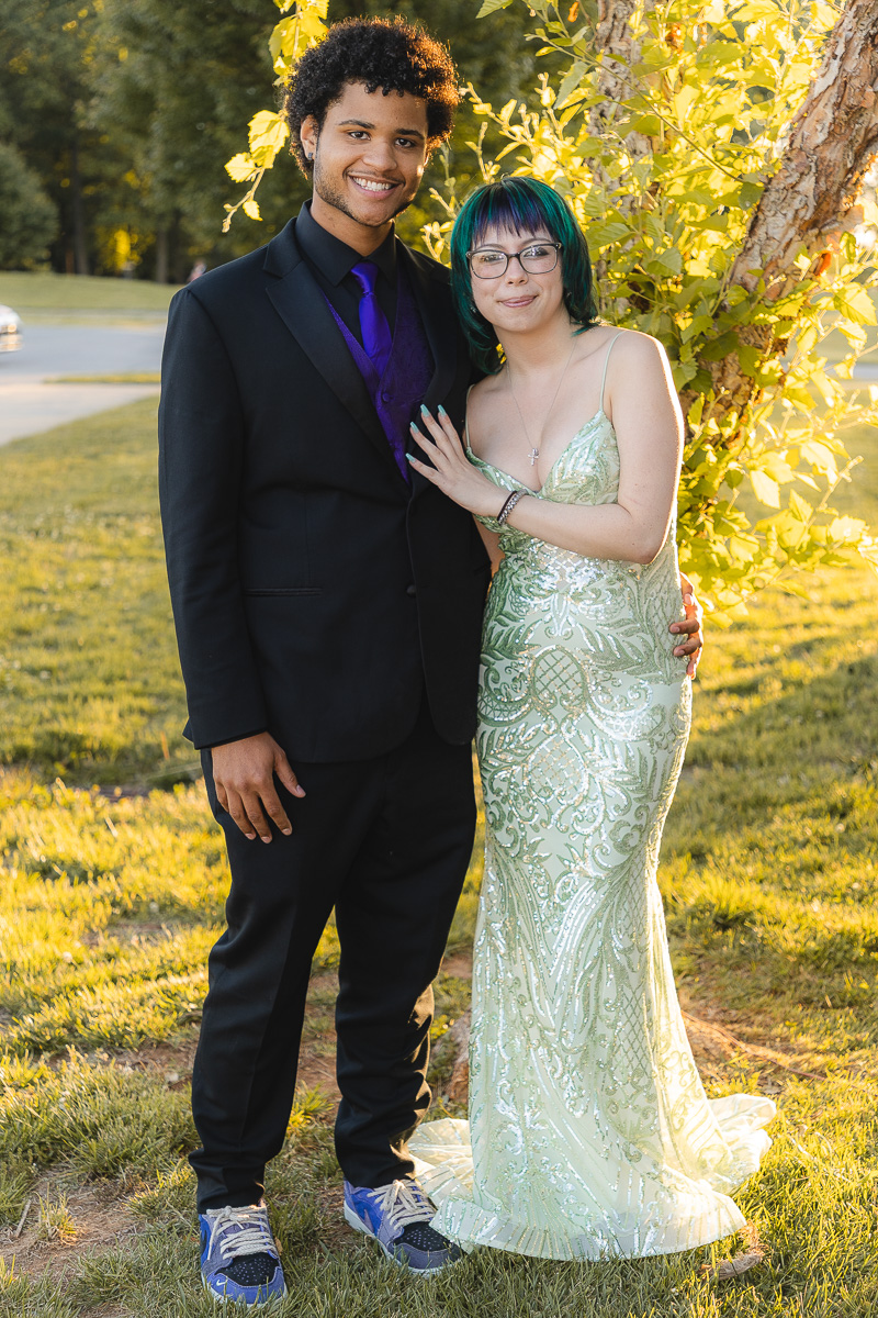 Couple posing outdoors in formal attire: man in a black suit with a purple tie and sneakers, woman in a mint green sequined gown with teal hair and glasses, standing beside a tree in sunlight.