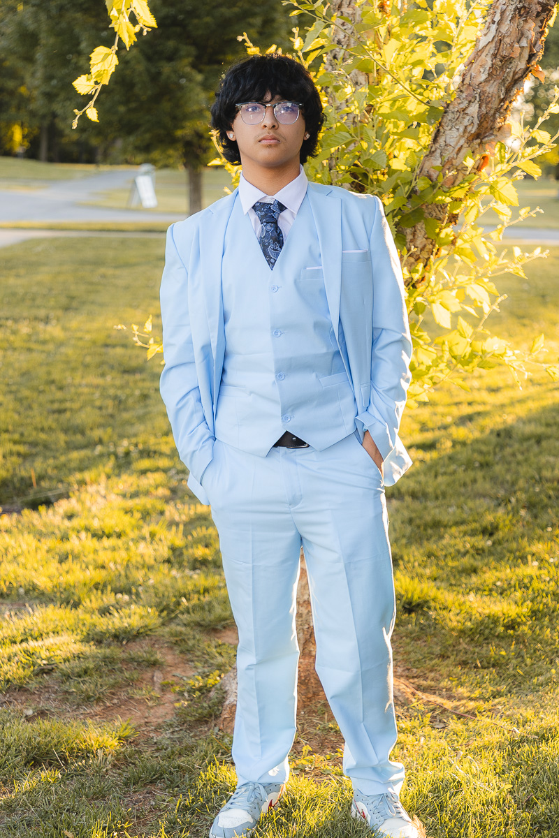 Man in a light blue suit and tie stands outdoors by a tree, hands in pockets, wearing glasses.