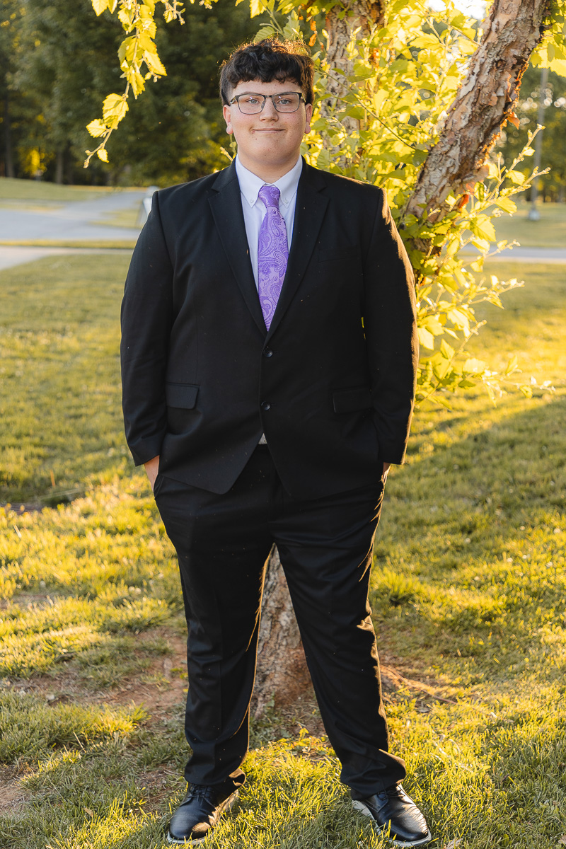 Young man in a black suit with a purple tie stands by a tree in a park during golden hour, hands in pockets.