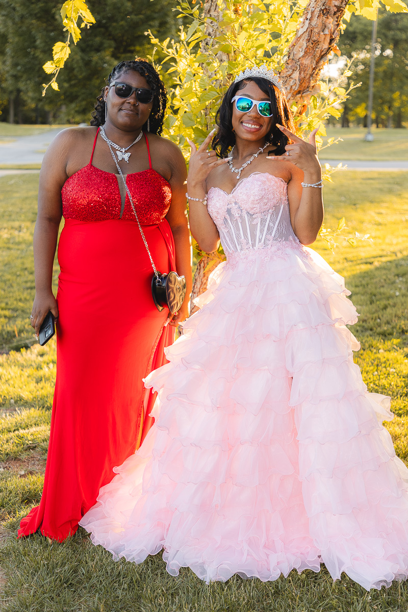 Two women in formal gowns posing outdoors by a tree; one in a red sequin dress, the other in a pink ball gown with a tiara and sunglasses on.