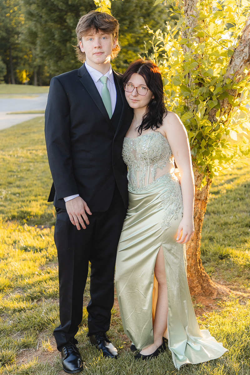 Couple dressed for a formal event posing outdoors beside a tree in warm sunlight; man in a black suit and mint tie, woman in a pale green gown with lace detailing.
