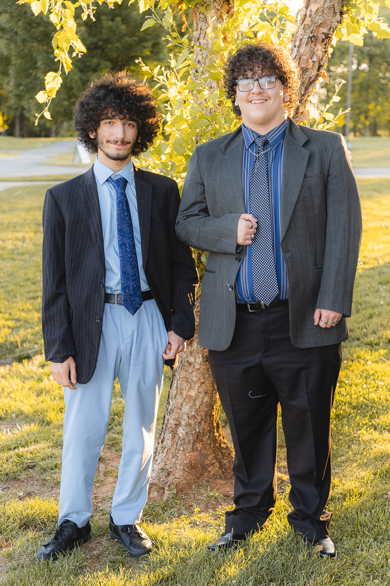 Two young men in suits standing beside a tree in a sunny park, holding hands and smiling at the camera.