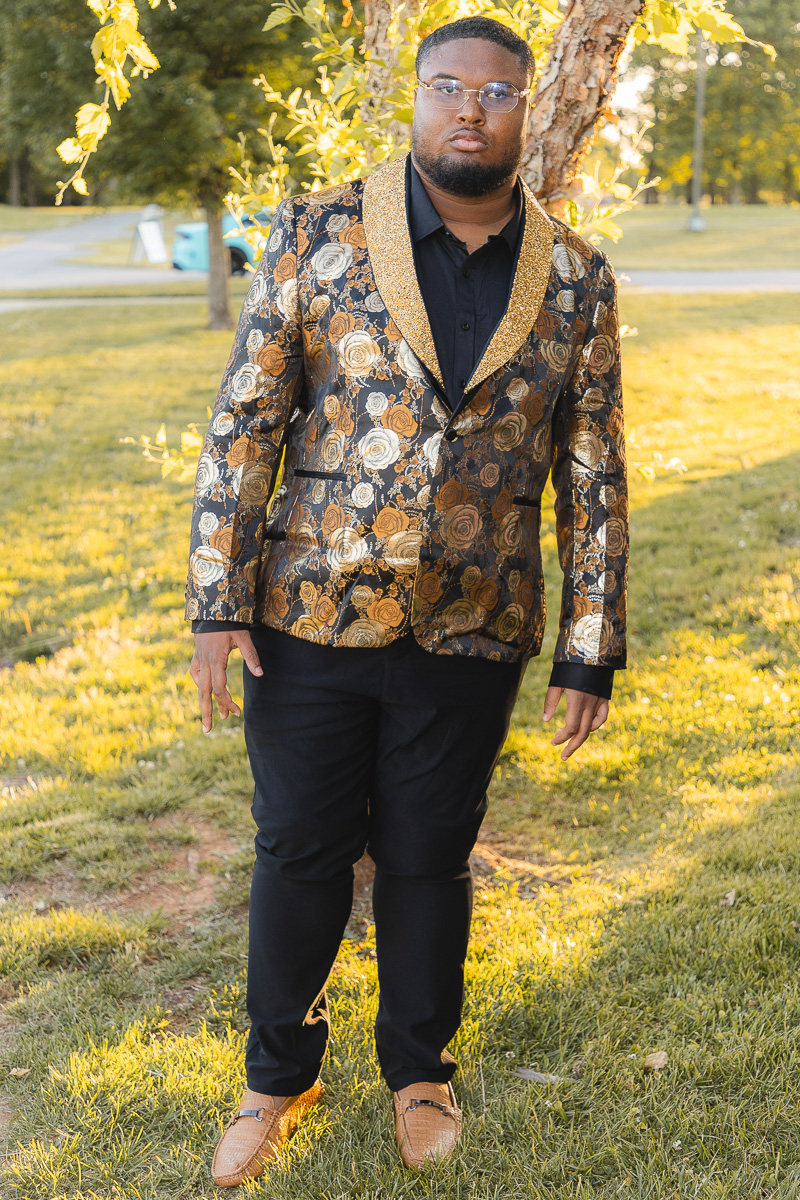 Man posing in a gold-rose patterned blazer, black shirt, and glasses in a sunlit park.