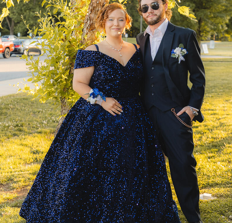 Couple in formal wear posing by a tree on a sunny lawn; woman in a navy blue sequin ball gown with corsage, man in a black suit with boutonniere.