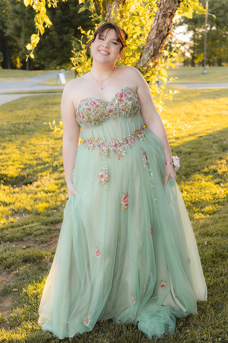 Young woman in a mint green floral-embellished ball gown standing in a sunlit park, smiling at the camera.