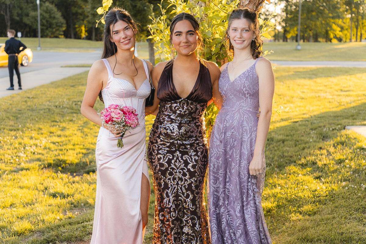 Three young women in formal gowns pose together on a sunny park lawn, one holding a pink bouquet.