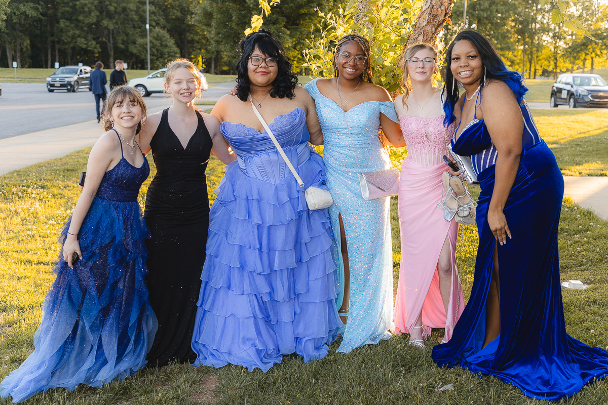 Six young women in colorful formal gowns pose together on a sunny grassy area, arms around each other.