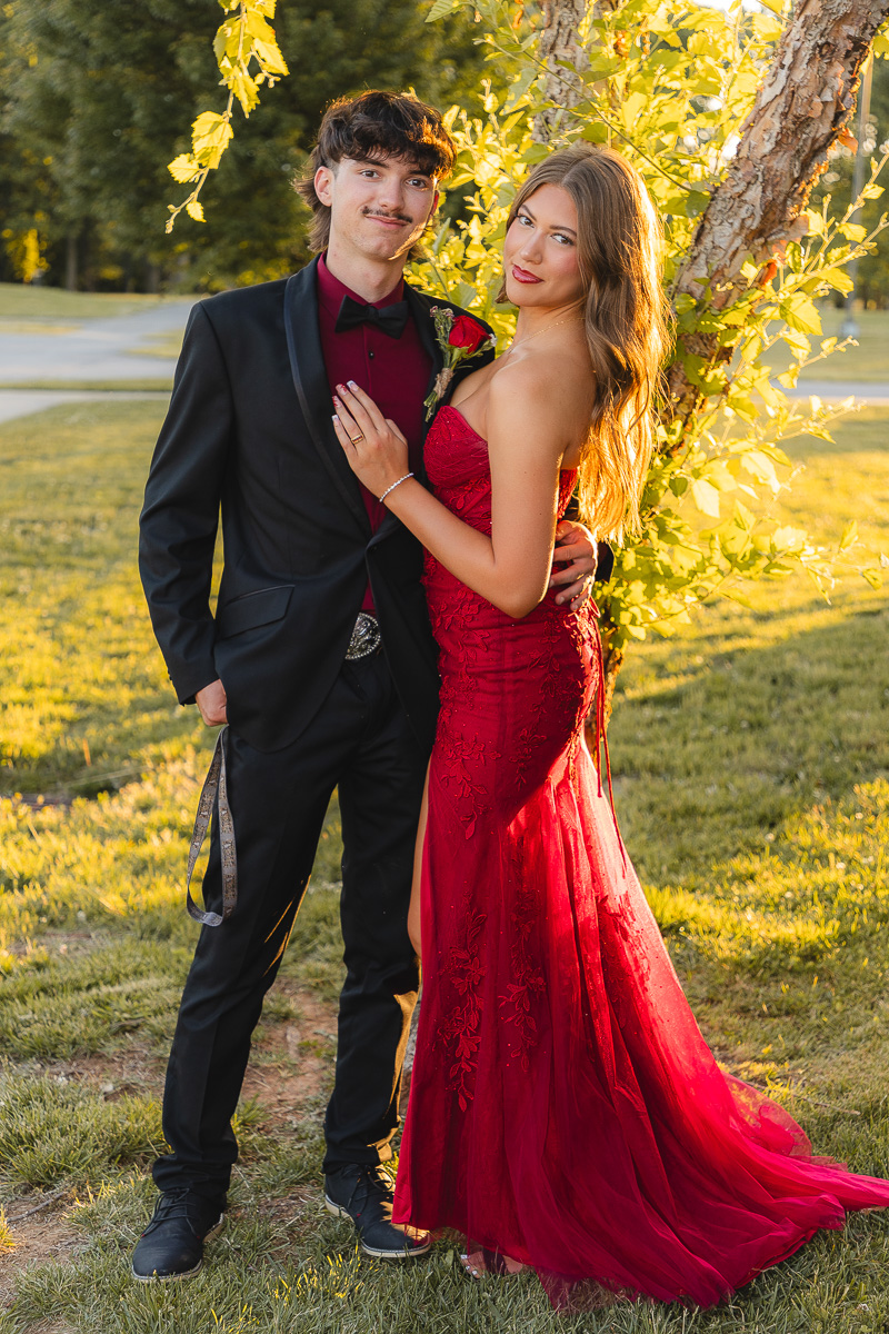 Couple in formal wear posing outdoors by a sunlit tree; woman in a red lace gown and man in a black suit with burgundy shirt.