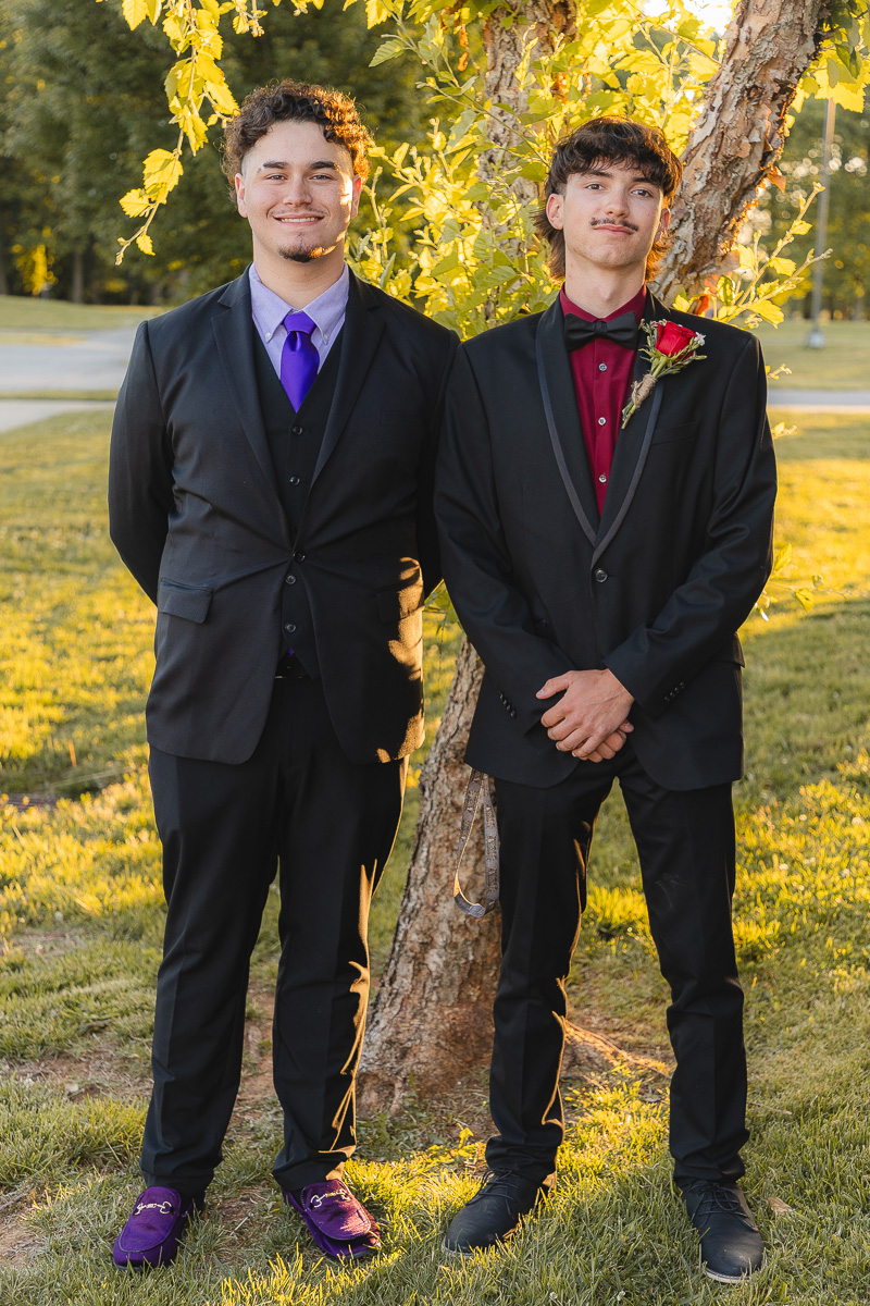 Two young men in black tuxedos pose beside a tree in a sunlit park, smiling at the camera; one wears a purple tie and shoes, the other a red shirt with a boutonniere.