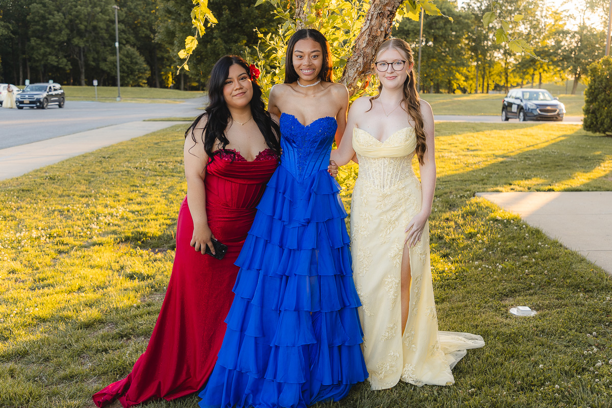 Three young women in formal gowns stand together on a grassy park at sunset, wearing red, blue, and pale yellow dresses and smiling at the camera.