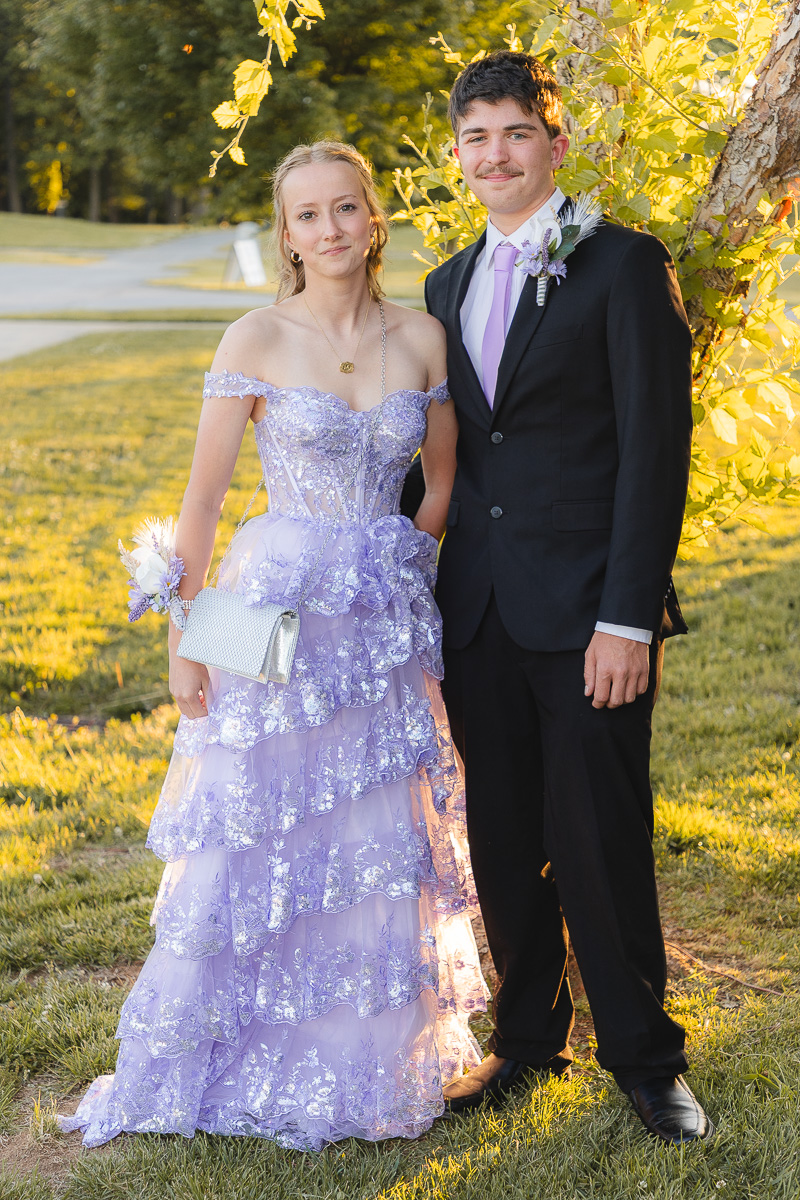 Couple posing outdoors in prom attire: girl in lavender ruffled gown with a silver clutch, boy in a black suit with lavender tie by a sunlit tree base