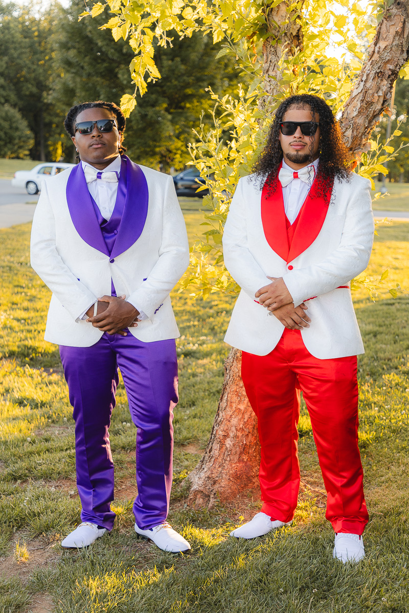 Two men in white suits with bold purple and red vests, bow ties, and sunglasses, posing outdoors beside a tree.