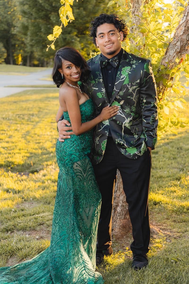 Couple posing outdoors in formal wear; woman in a green embellished gown and man in a black suit with a green floral pattern, standing by a tree.