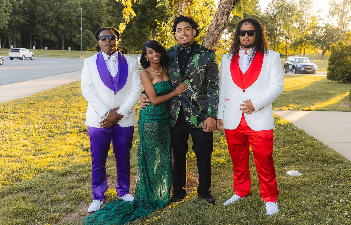 Four friends pose outdoors in formal attire: two men in white jackets (one with purple accents, the other with red) flanking a woman in a green sequined gown and a man in a black-and-green floral blazer beneath a tree.