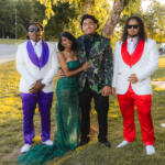 Four friends pose outdoors in formal attire: two men in white jackets (one with purple accents, the other with red) flanking a woman in a green sequined gown and a man in a black-and-green floral blazer beneath a tree.