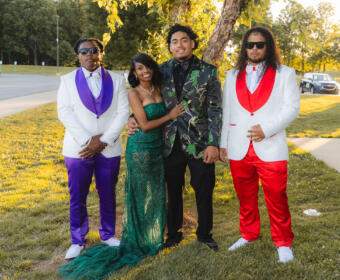 Four friends pose outdoors in formal attire: two men in white jackets (one with purple accents, the other with red) flanking a woman in a green sequined gown and a man in a black-and-green floral blazer beneath a tree.