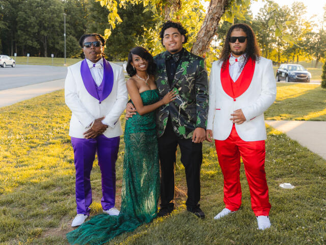 Four friends pose outdoors in formal attire: two men in white jackets (one with purple accents, the other with red) flanking a woman in a green sequined gown and a man in a black-and-green floral blazer beneath a tree.