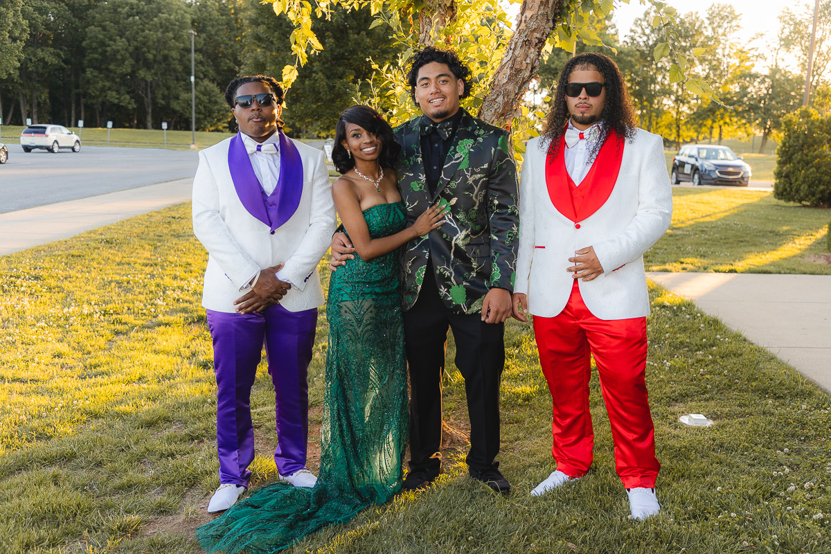 Four friends pose outdoors in formal attire: two men in white jackets (one with purple accents, the other with red) flanking a woman in a green sequined gown and a man in a black-and-green floral blazer beneath a tree.