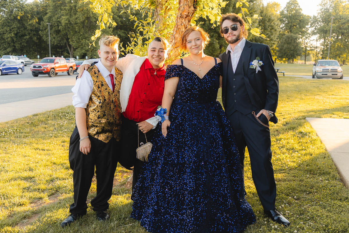 Four friends pose together outdoors under a tree, dressed for a formal event: two men in suits (one in a gold-vested outfit, one in a black suit) and a woman in a sparkling blue gown with a corsage, with sunlight lighting the scene.
