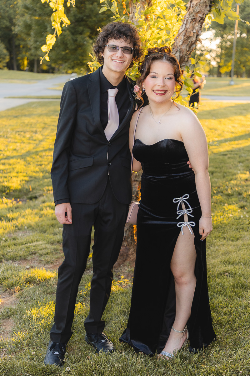 Smiling couple in formal wear posing outdoors: man in a black suit with a pale pink tie, woman in a black velvet gown with side lace ties, by a tree in a park at sunset