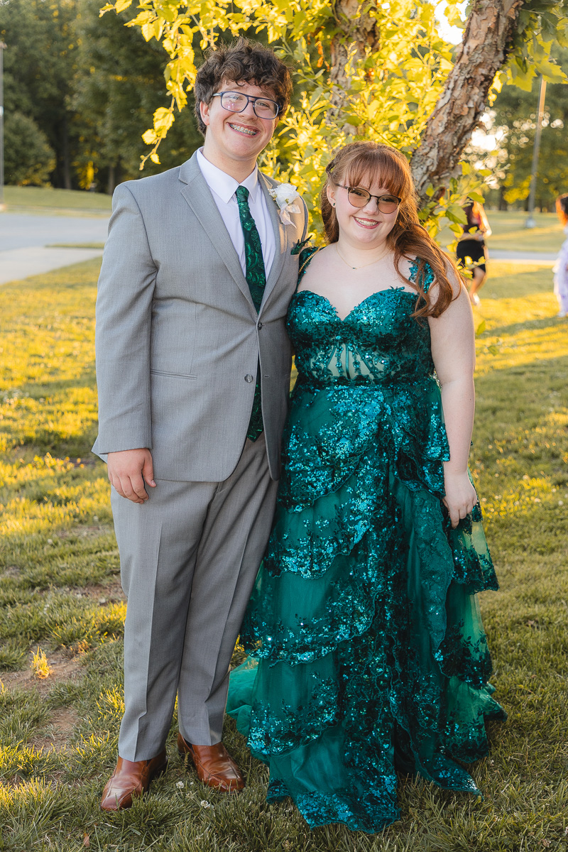 Two friends in formal wear posing outdoors: man in a gray suit and green tie, woman in a teal sequined gown, both smiling under a tree.