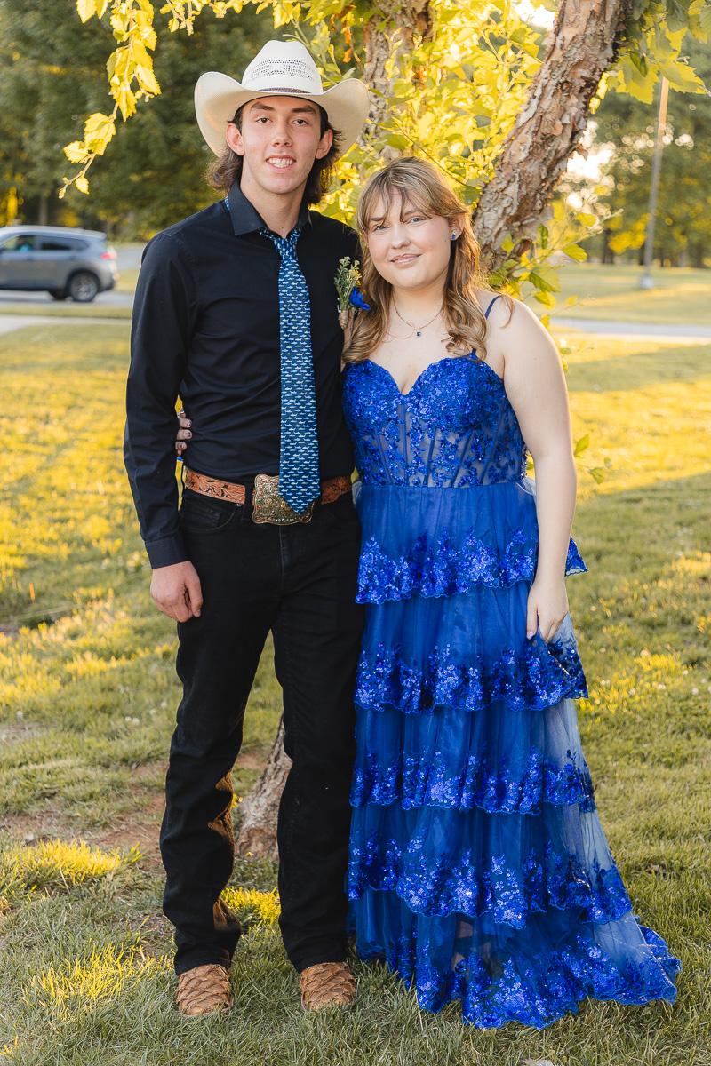 Young couple posing outdoors in a park; man wears a black shirt, blue patterned tie, cowboy hat, and belt, with a woman in a blue sequined gown beside him under a tree.