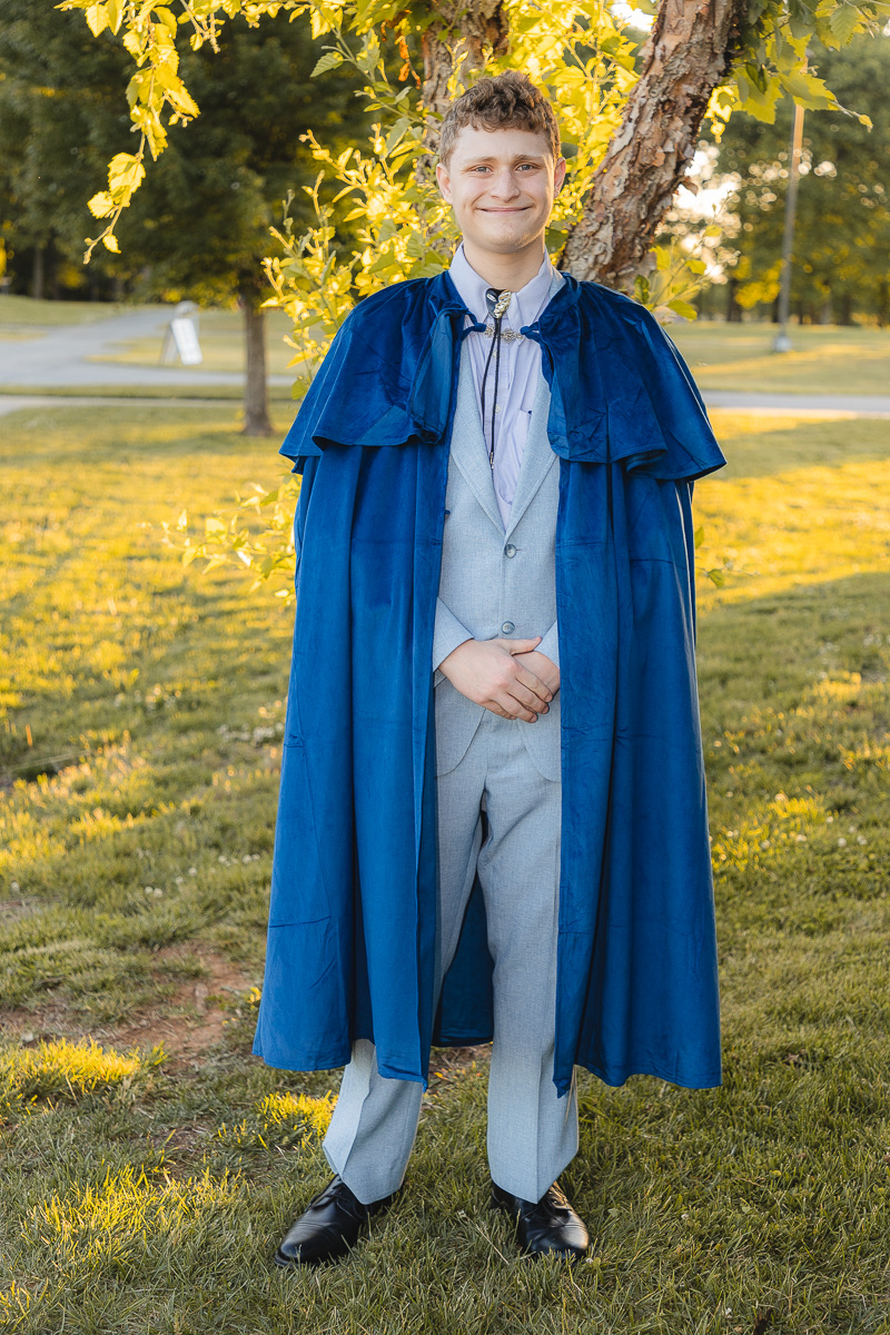 Smiling graduate in a blue gown over a gray suit, standing on a sunlit park lawn with trees nearby.