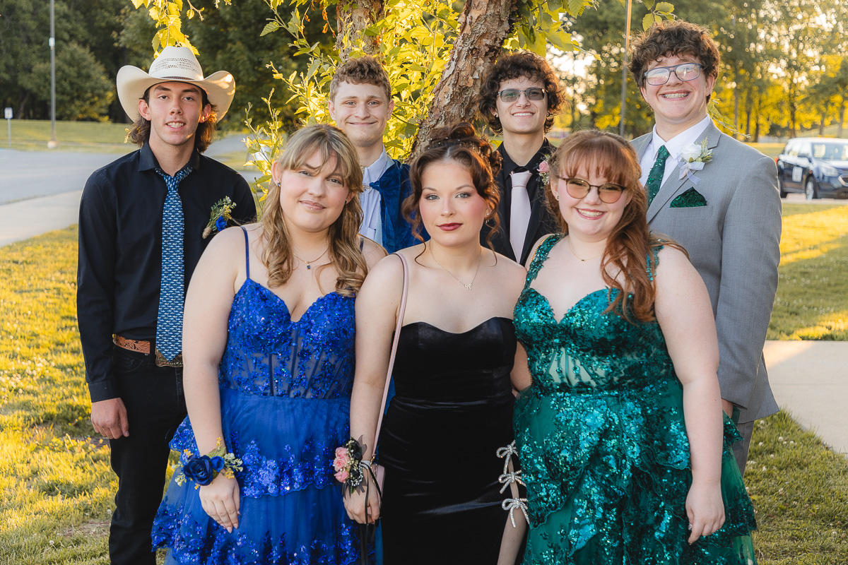 Six teenagers in formal attire pose together outdoors, three girls in colorful dresses and three boys in suits beneath a tree sunlit yard.