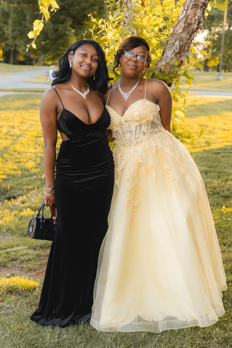 Two young women in formal gowns posing together in a park, one in a black velvet dress and the other in a pale yellow gown, smiling by a tree in autumn light.