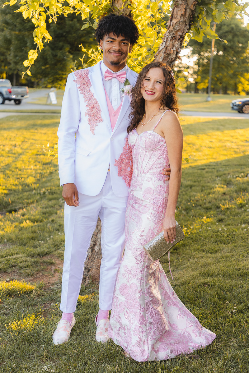 Young couple in pink and white formal attire posing together outdoors under a tree.