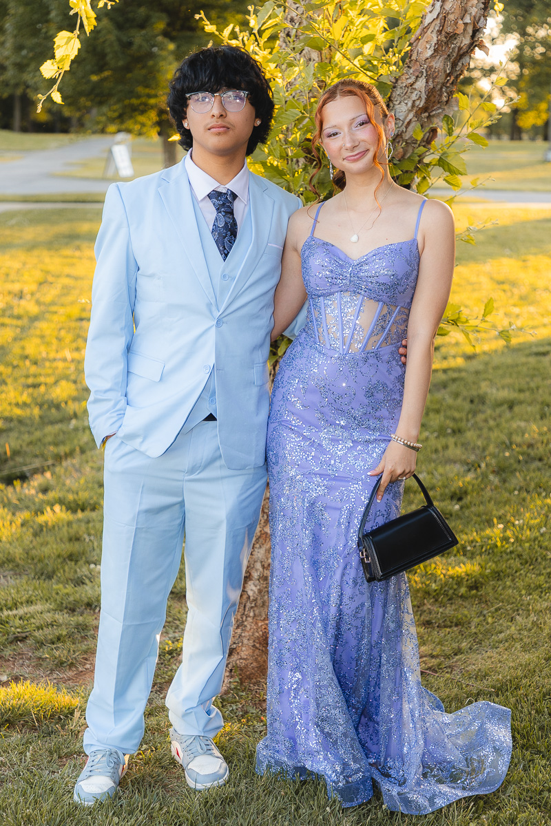 Young couple posing outdoors in formal wear: man in a light blue suit and tie, woman in a lavender sequined gown with a black clutch.