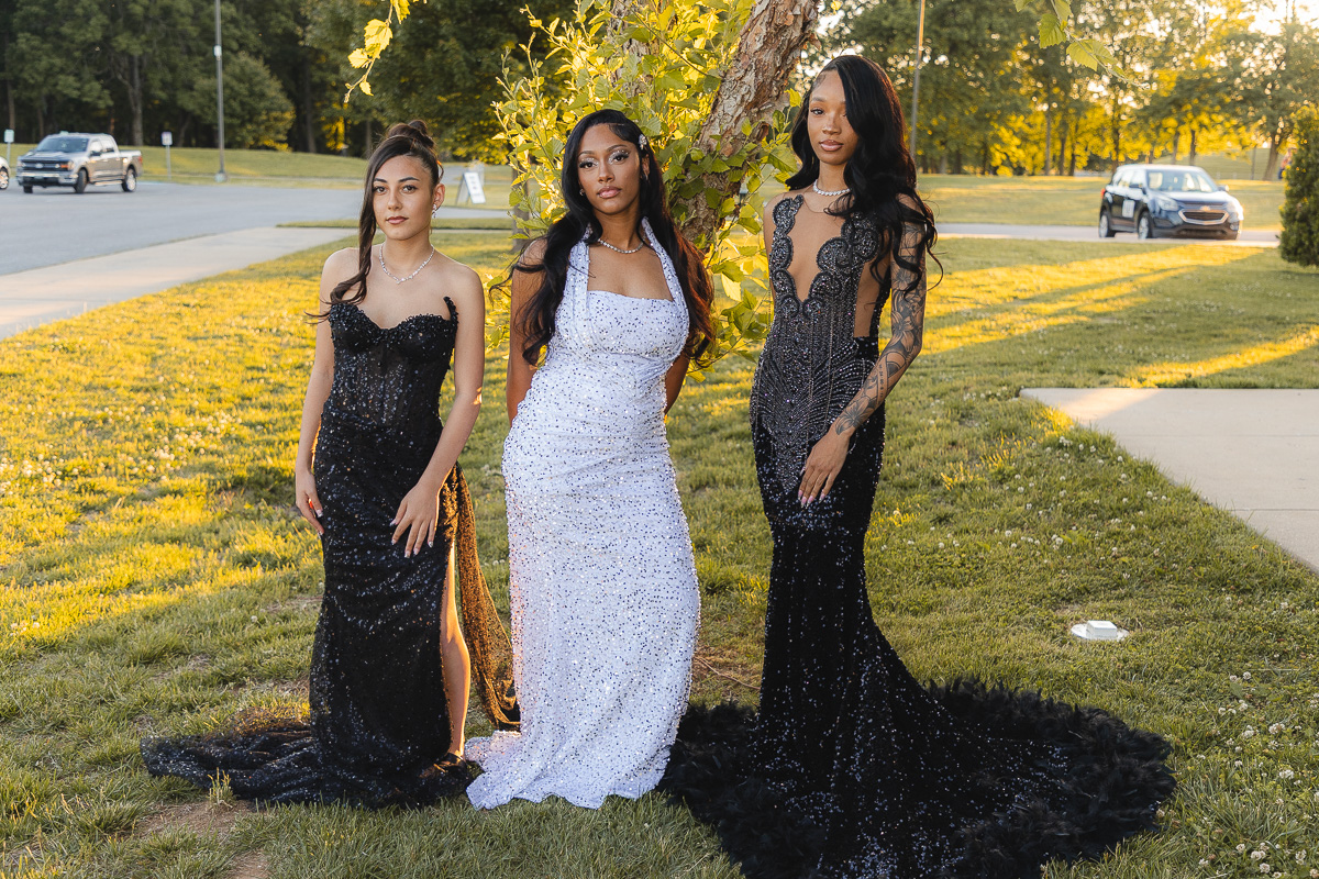 Three women in formal gowns pose together outdoors near a tree at golden hour. Two wear black gowns, one wears a white beaded dress, all looking at the camera.