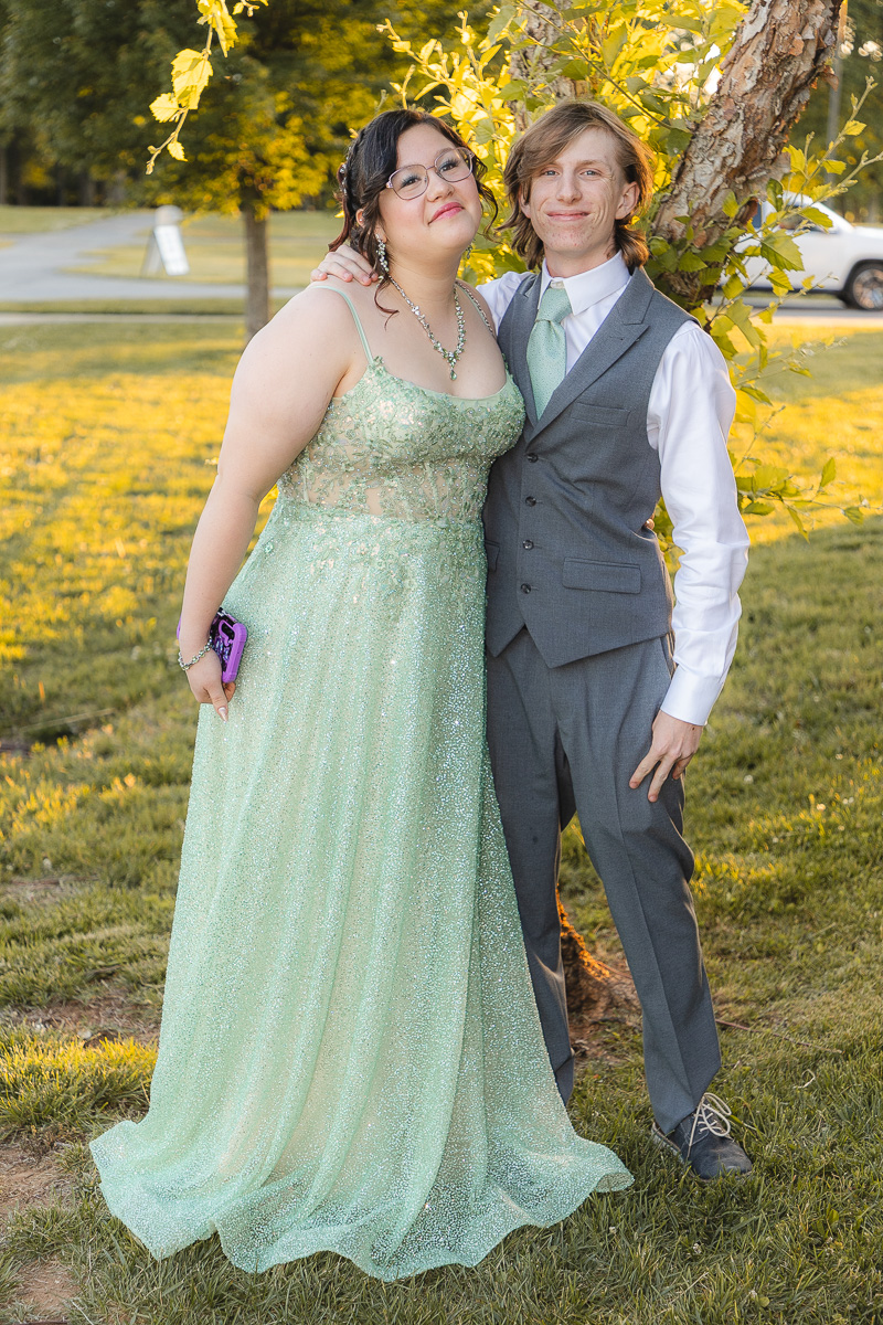 Two teens in formal wear posing outdoors: girl in a mint green sparkly gown, boy in a gray suit with a mint tie, autumn leaves in the background.