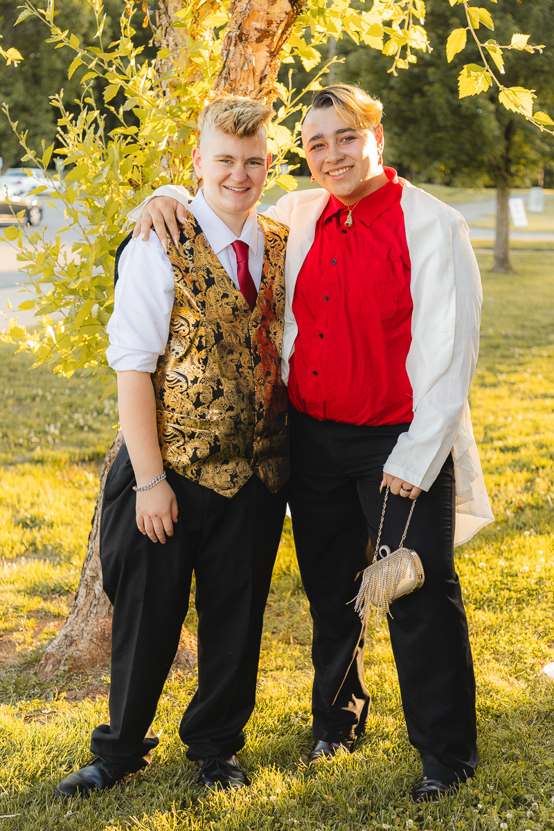 Two young people pose together outdoors in formal outfits by a tree, smiling at the camera.