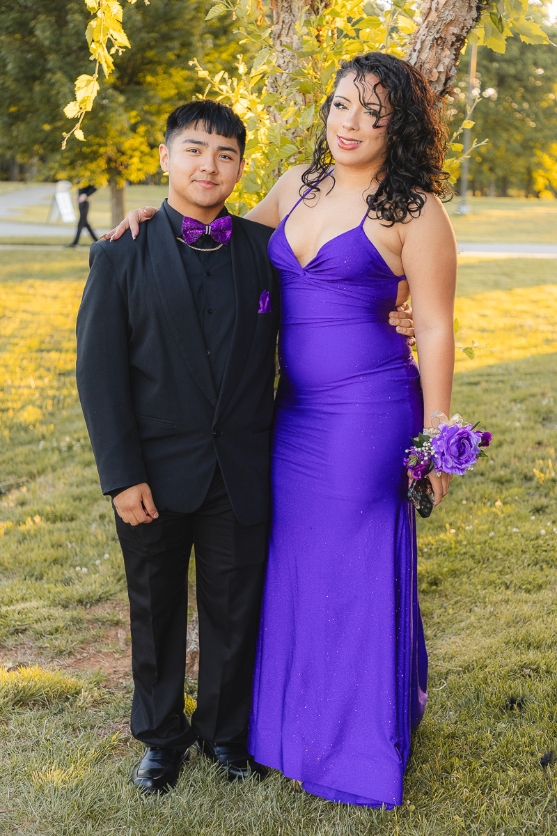 Couple posing outdoors in formal attire: man in black suit with purple bow tie and woman in a purple gown holding a matching corsage.