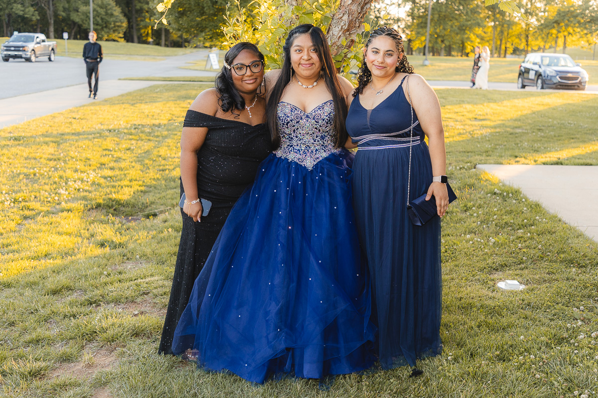 Three young women in formal gowns pose together under a tree on a sunny day: left in an off‑the‑shoulder black dress, center in a royal blue embellished ball gown, right in a navy dress with a small purse.