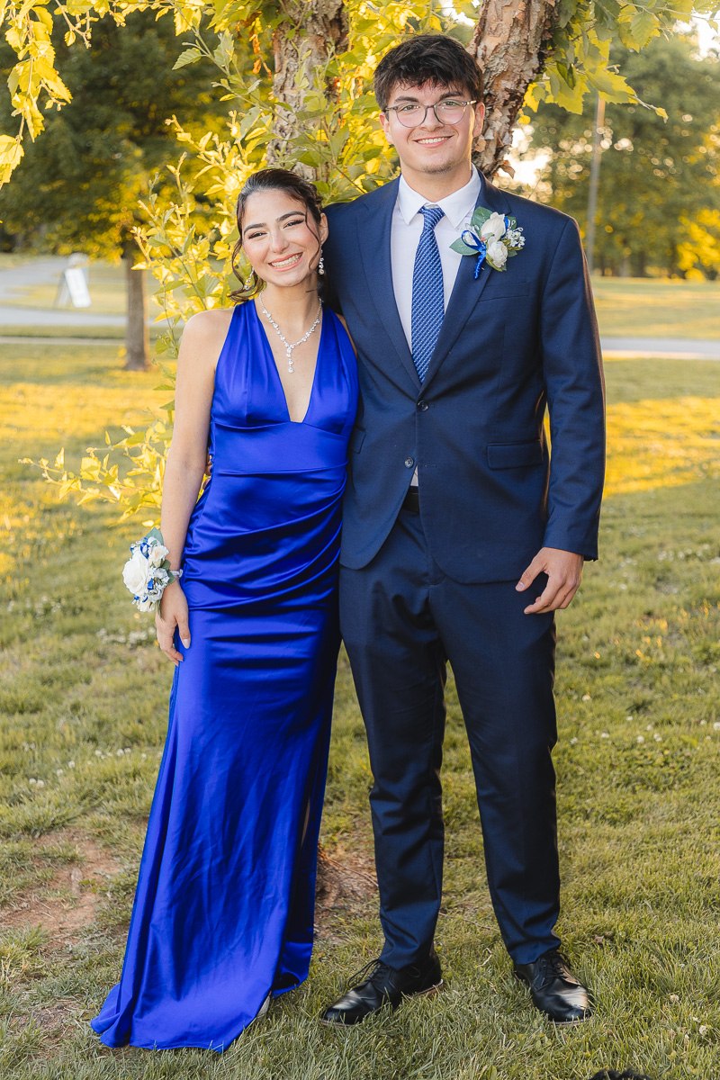 Two friends posing outdoors in formal wear: a woman in a royal blue gown with a corsage and pearl jewelry, and a man in a navy suit with a boutonniere, standing beside a tree on a sunny lawn.