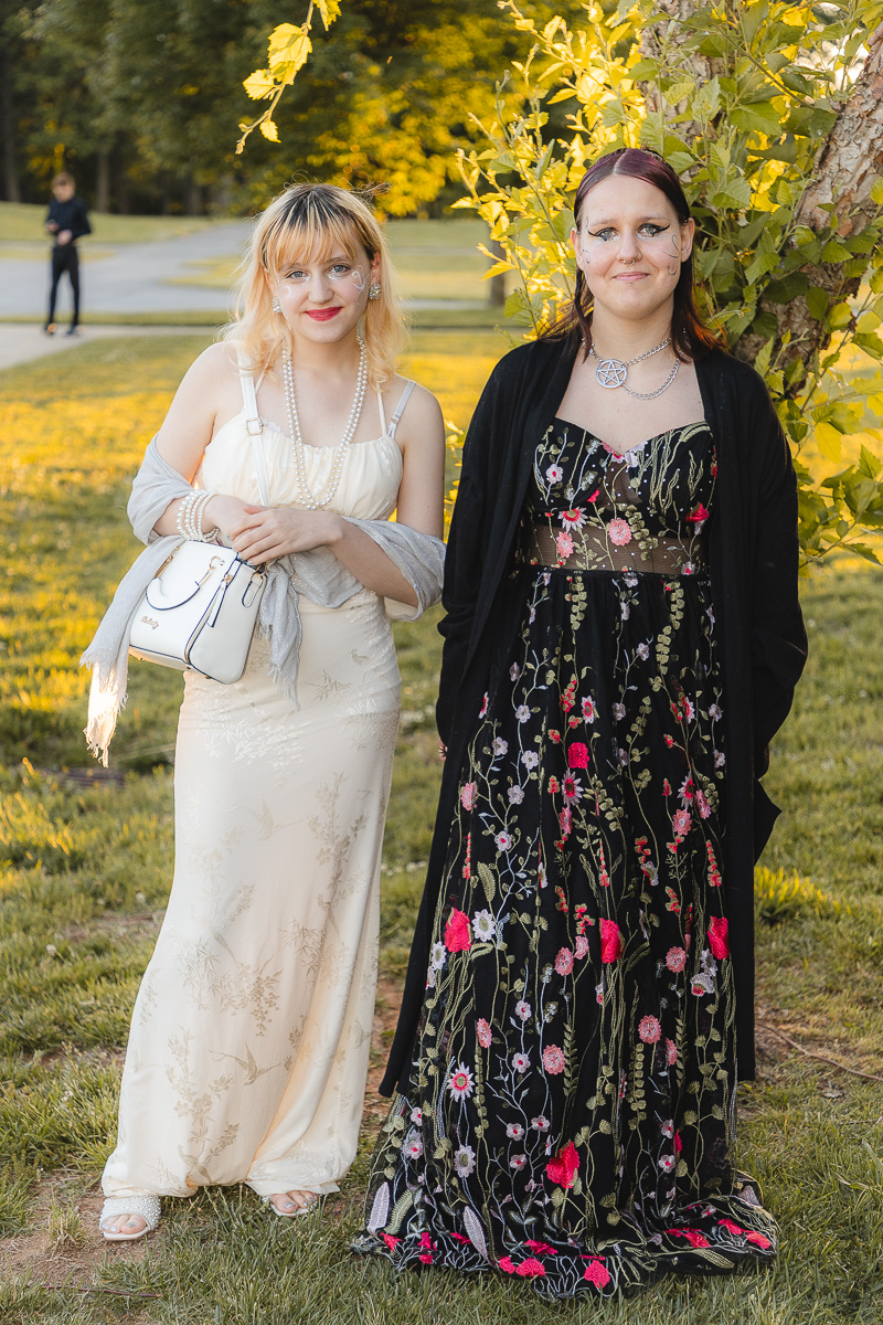 Two women in formal dresses pose outdoors in a park, one in a cream gown with pearls and a white handbag, the other in a black floral gown with a cardigan by a tree.