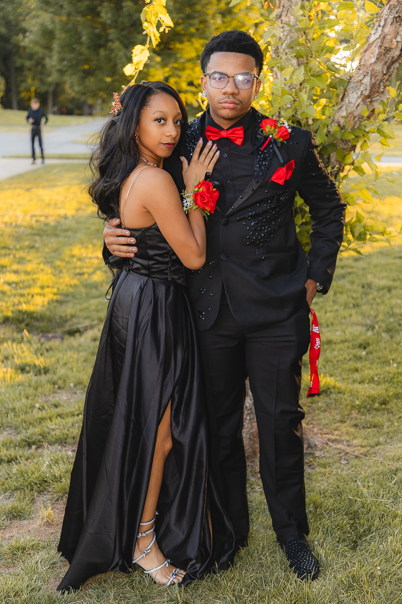 Couple in formal black outfits posing by a tree outdoors; woman in a black gown with slit and heels, man in a black suit with red bow tie and boutonniere.
