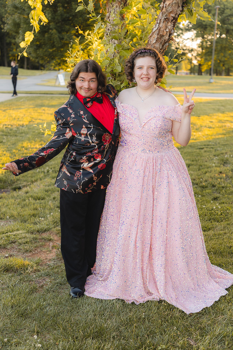 Two teens in formal wear pose together in a sunlit park, the girl in a pink glittery gown making a peace sign while the boy wears a floral jacket and red shirt smile.