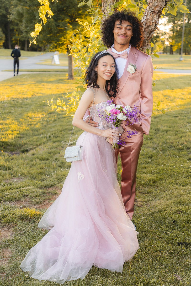 Couple in formal wear posing outdoors; woman in pink gown with bouquet, man in pink suit.