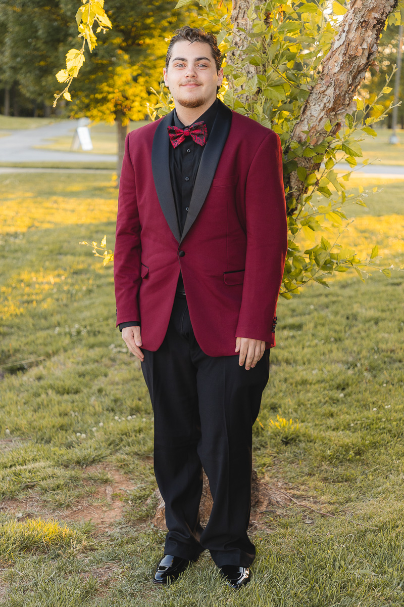 Young man in a burgundy tuxedo jacket with black lapels and a red bow tie stands beside a tree in a park, autumn setting.
