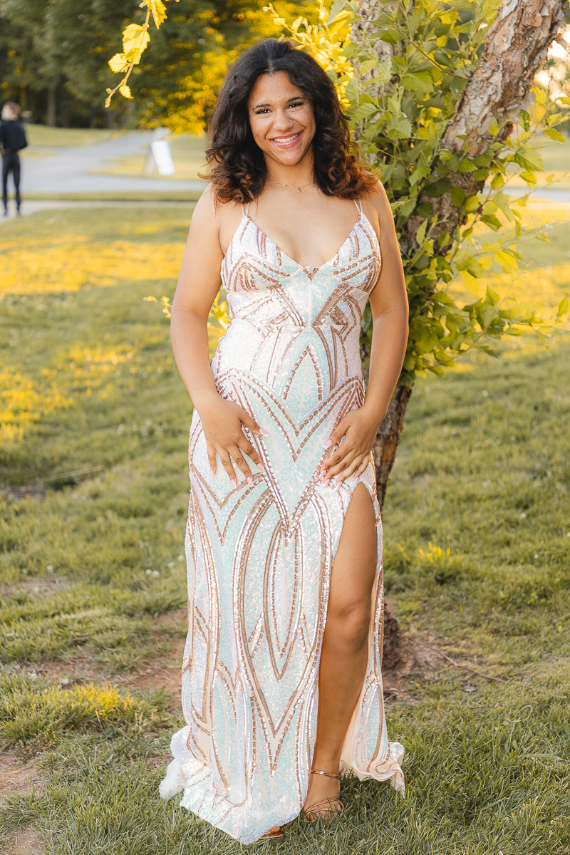 Smiling woman in a long, patterned gown standing beside a tree in a sunny park.