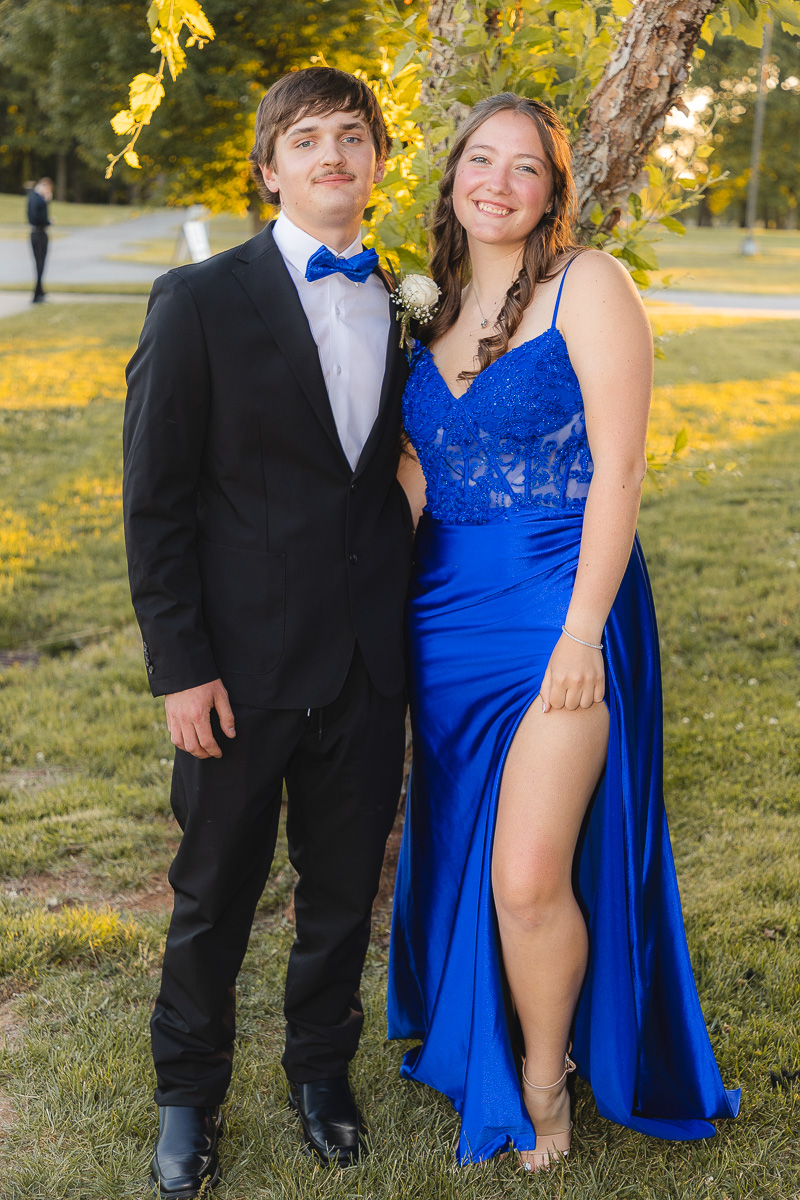 Young couple in formal wear posing outdoors; man in a black suit with a blue bow tie, woman in a blue lace gown with a high slit.