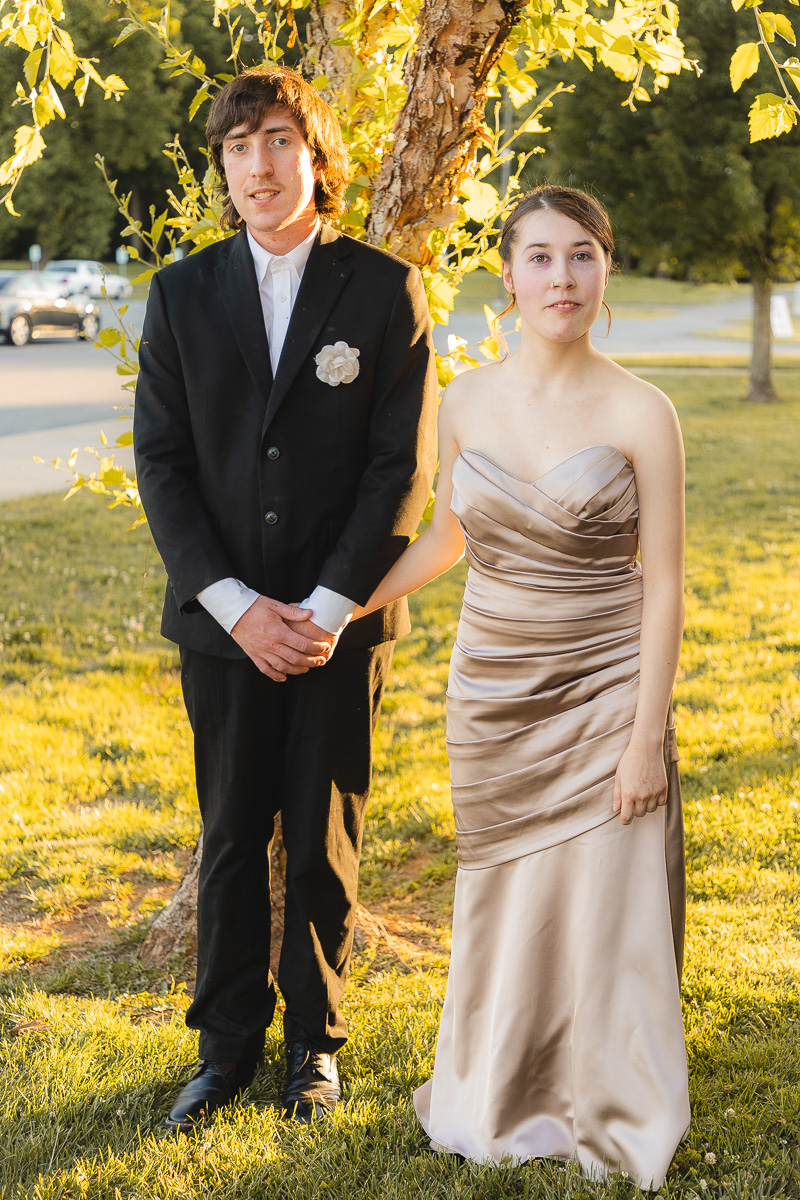 Young couple in formal attire outdoors; man in a black suit with a white boutonniere, woman in a champagne strapless gown, holding hands.