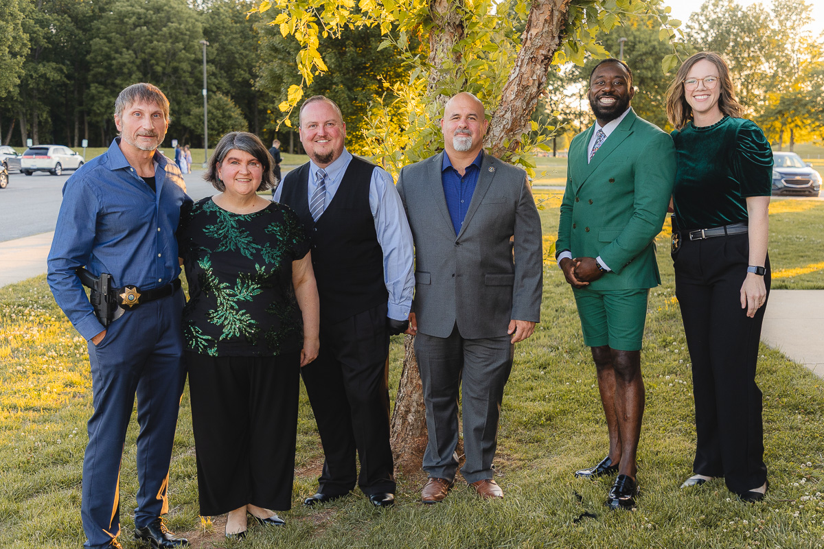 Six adults stand in a line outdoors by a tree, smiling for a group photo in daytime attire ranging from casual to formal.