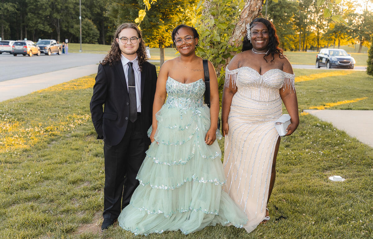 Three friends in formal attire posing outdoors by a tree, two women in gowns and a man in a suit, smiling at the camera.