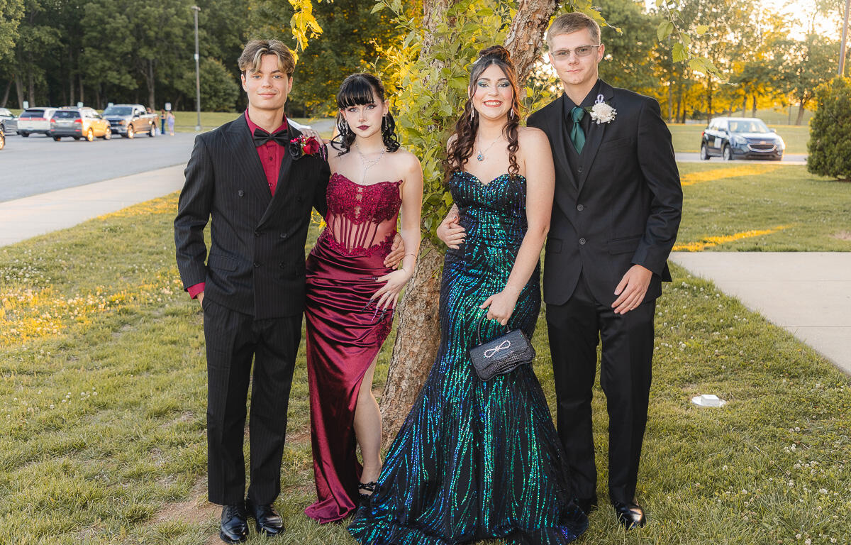 Four teens in formal wear posing by a tree on a sunny lawn—two girls in evening gowns and two boys in suits, smiling for the camera.