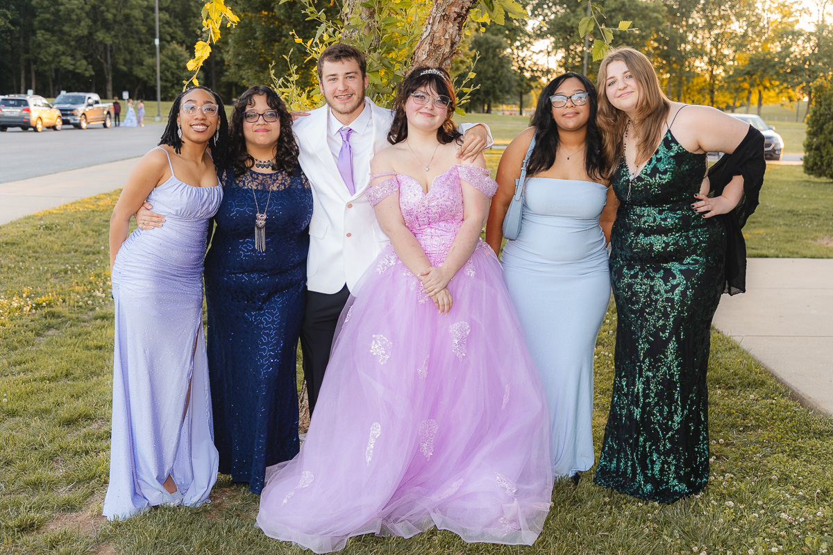 Six friends in formal wear pose outdoors for a group prom photo, central couple with lavender gown and white suit, sunset lighting behind them.
